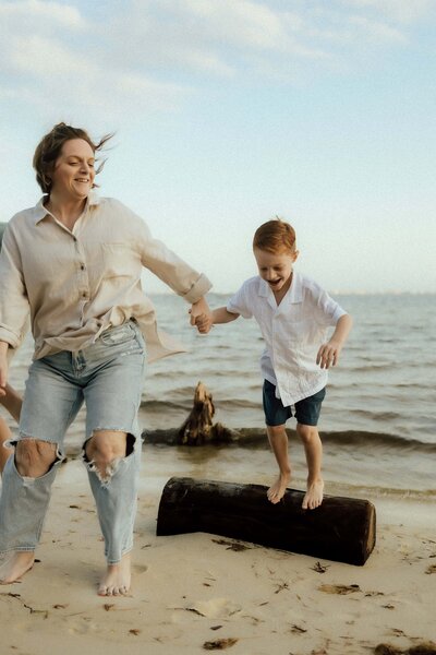 An adult and a child holding hands on a sandy beach; the child is jumping off a log while the adult smiles. The ocean and sky are in the background.