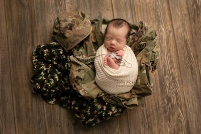A sweet newborn boy wrapped in soft fabric, peacefully laying on his dad's army clothes, creating a heartfelt and special moment, captured by Chunky Monkey Photography.