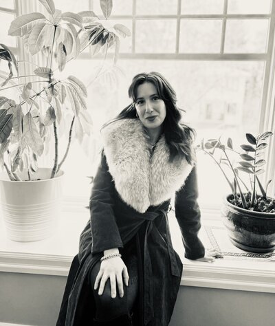A close up shot of Hilary Rota, posed in front of her computer, with her hand under her chin, smiling and wearing glasses