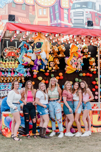 Rachel B Photography's Senior Rep Team of seven girl pose in front of a carnival game booth smiling at the camera.