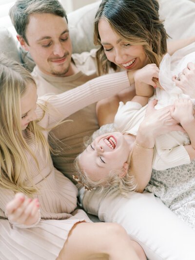 Mother, father, and 2 young, blonde daughters tickling each other and laughing sitting on a couch in their home