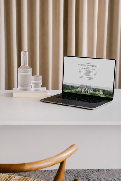 A desk with a laptop, water glass and cup. There's a curtain in the background and a chair pulled up to the table. The laptop displays a Showit website with SEO copywriting.