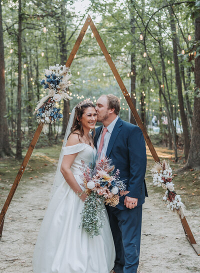 A happy wedding couple smiles and shares a loving moment beneath a rustic wooden triangle arbor decorated with blue and white flowers and string lights in an outdoor setting, captured by Quinn Photography LLC.