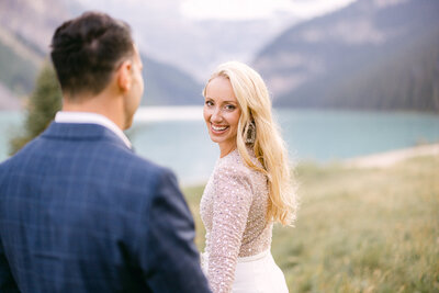 This joyful photograph captures the bride looking back at the camera, laughing as she holds the groom's hand while they walk away from the Fairmont Chateau Lake Louise Hotel. With the stunning Lake Louise ahead, the moment perfectly blends happiness and the beauty of their surroundings.