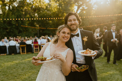 husband and wife smile with plates of food at wedding reception
