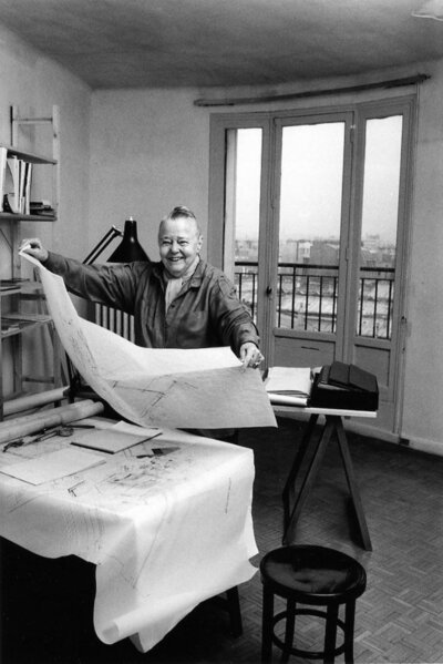 Charlotte Perriand in 1991, standing next to a table filled with design supplies and sketches, holding a piece of paper showcasing her innovative design ideas.