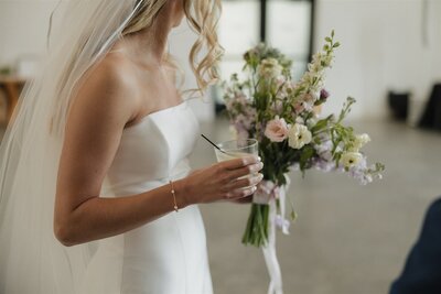 Bride holds spring bridal bouquet and a cocktail as she talks to a guest during happy hour at her wedding