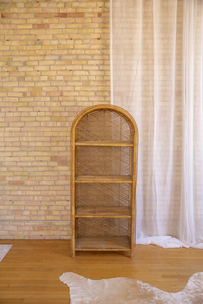 A rattan shelf with four shelves standing up in front of a brick wall.