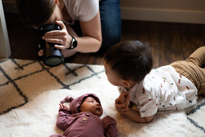 Photographer kneeling on the ground getting a close up photo of a baby wearing a light pink outfit laying on a white rug next to her toddler brother for a lifestyle newborn photo shoot