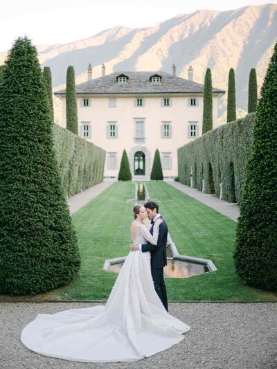 Bride and groom embracing in front of an Italian villa at Villa Balbiano, Lake Como, framed by manicured cypress trees — Portfolio Thomas Raboteur Wedding Photographer.