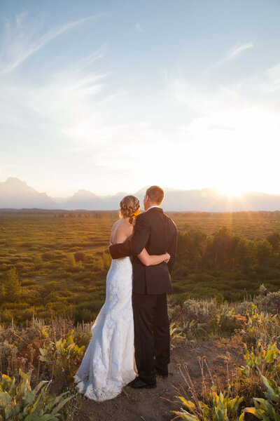 A bride and groom at sunset at Schwabacher’s Landing in Grand Teton National Park. The bride’s elegant satin wedding dress features an off-the-shoulder design and gold embroidery, while the groom wears a tailored burgundy suit. The reflection of the Tetons glows on the still water behind them.