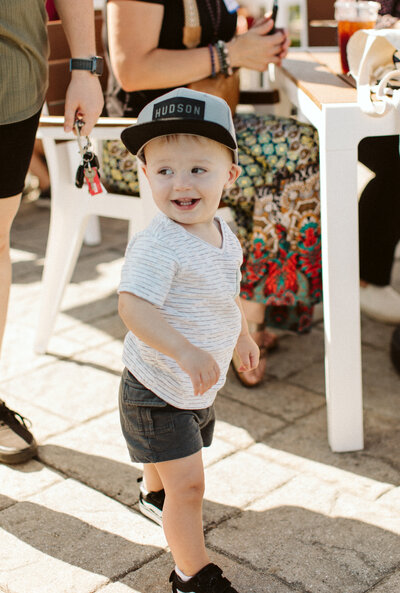 Child with a baseball cap smiling at someone behind him.