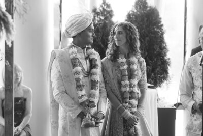 South Asian bride and groom look at each other warmly during their ceremony, wearing traditional attire and floral garlands.