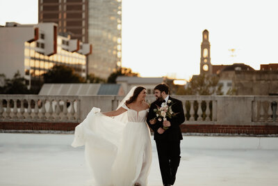 Bride and groom walking on a rooftop in downtown Winston Salem at sunset while the bride holds her dress and veil.