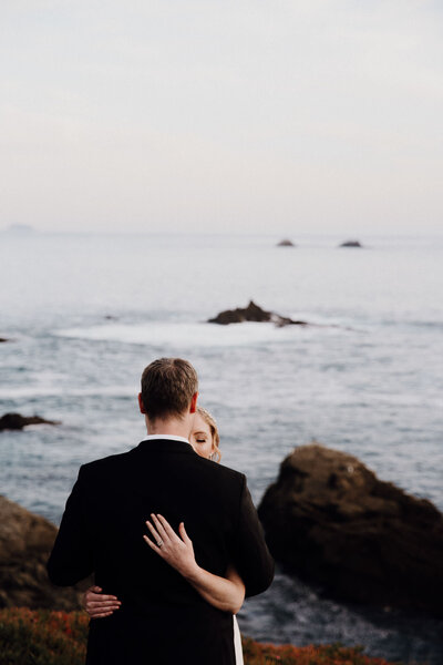 Bride wraps hands around groom and holds him tight. Groom  almost completely covers bride. Ocean and rocks in background
