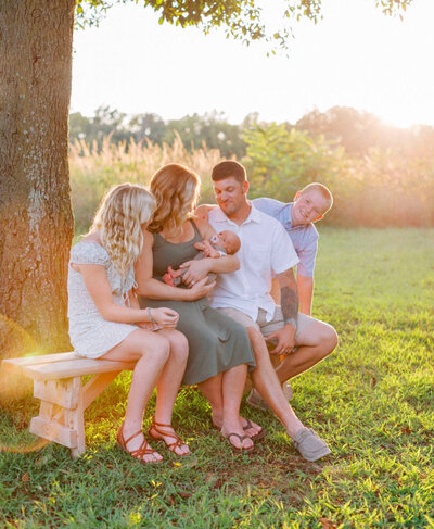 A mother holding her newborn baby while her other two kids. and husband arre sitting next to her on a bench under a tree
