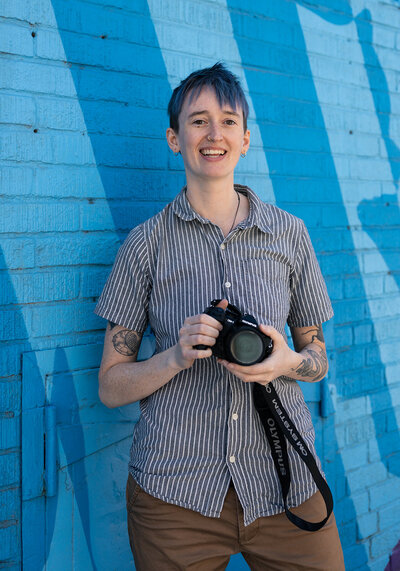 photographer River Dantzler smiles and holds up their camera. they have short blue hair and tattoos and wear a striped button down shirt.