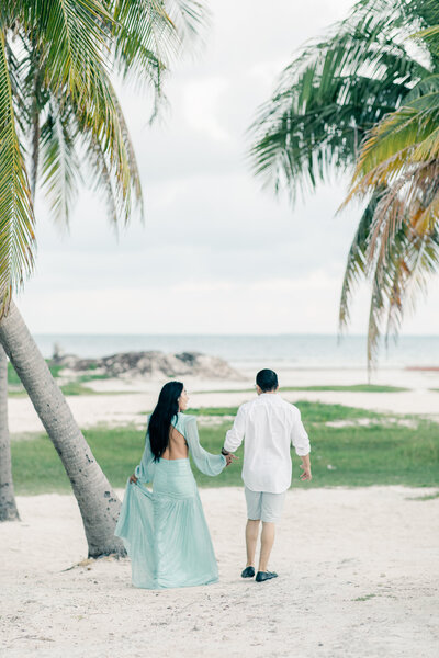 A girl wearing fA beautiful girl wearing a beautiful flowy Patbo sea foam teal green dress and a guy running holding hand with palm trees at beach