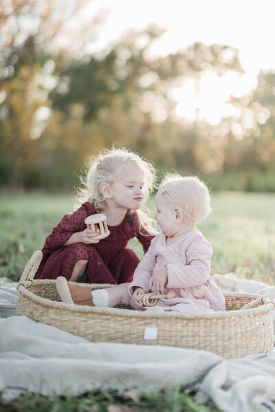 Young girl kissing her baby sibling in a woven basket during a cozy outdoor family photo session with a Kansas City family photographer.
