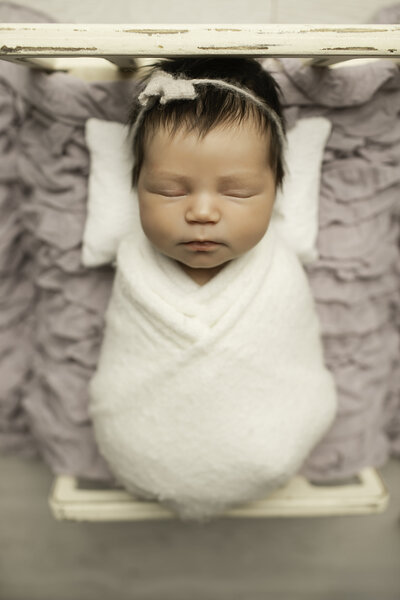 A beautiful newborn girl with a full head of hair, wrapped in a soft white fabric, peacefully resting on a cozy bed, captured in a serene and tender moment by Chunky Monkey Photography.