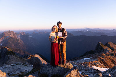 bride and groom are kissing at sunset wearing backpacks with signs saying they eloped on a peak. they have their hands in the air and face away from the camera
