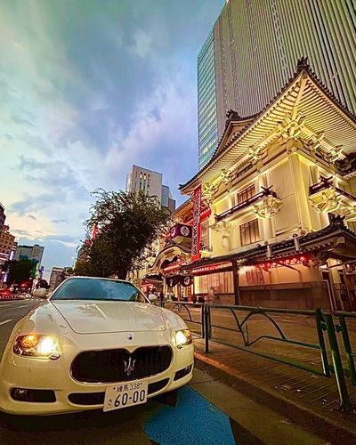 Luxury white Maserati parked on a vibrant Ginza street in Tokyo at dusk, surrounded by glowing storefronts and city life.