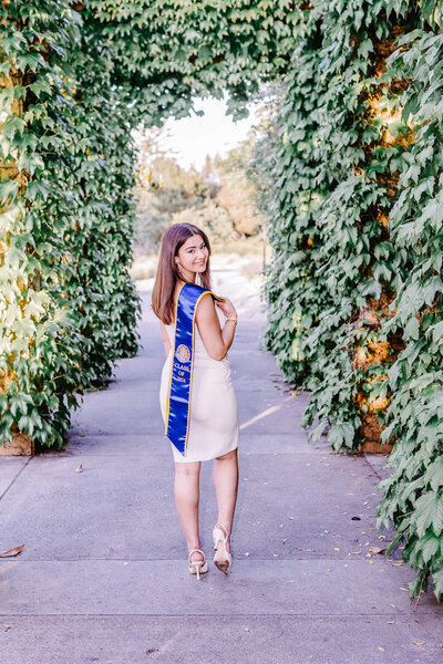 A brown haired young woman wearing a white dress and a UCI graduation sash standing with her hand on her hip in an alcove of vines
