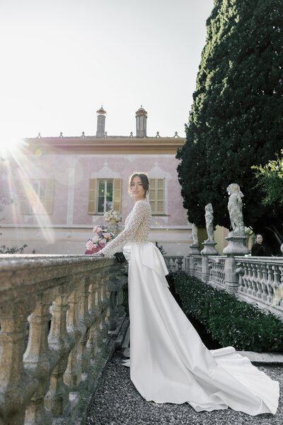 Bride in long-sleeved wedding gown standing on stone terrace at Villa Pizzo in Lake Como, Italy, with sun flare and romantic European architecture in the background
