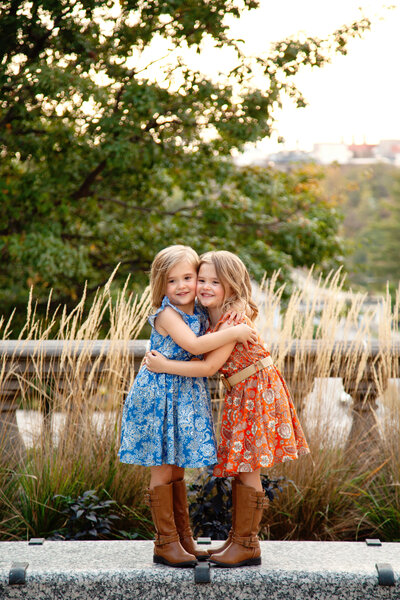 Young twin girls in dresses and tall boots hugging cheek to cheek and smiling.