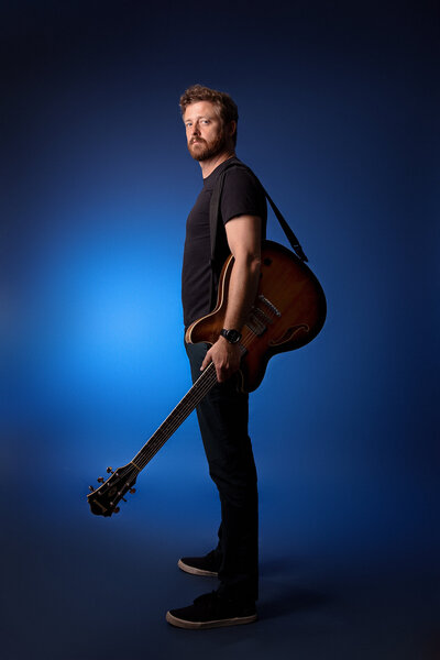 A man with long curly hair and a beard, dressed in a black button-up shirt, stands against a dark background. Perfect for branding photography Jacksonville, he holds a guitar strap over his shoulder, with a red electric guitar visible behind him.