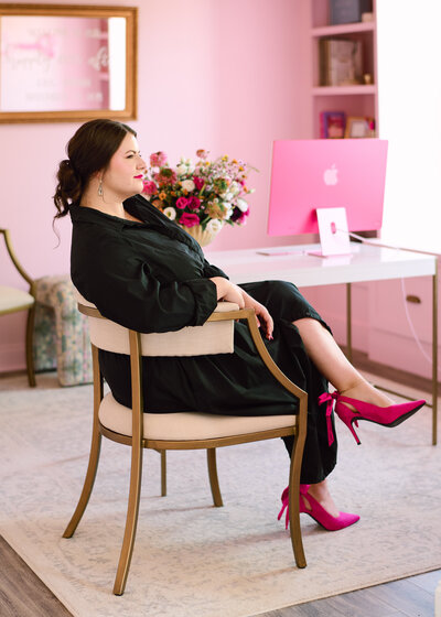 Brunette woman wearing black dress, sitting in front of desk looking towards windows