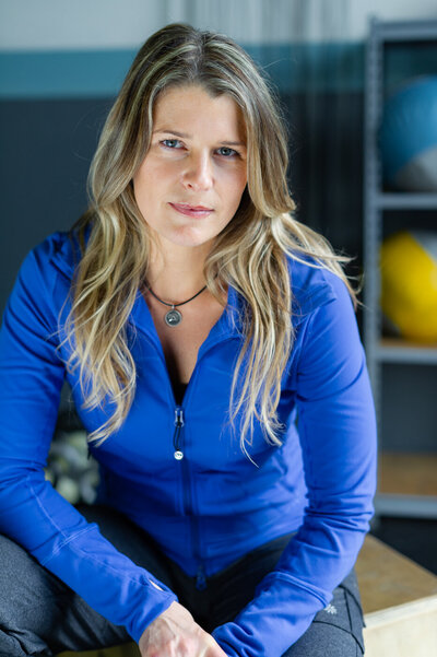 Fitness trainer headshot of a woman with long brown hair wearing a blue zip up sweatshirt.