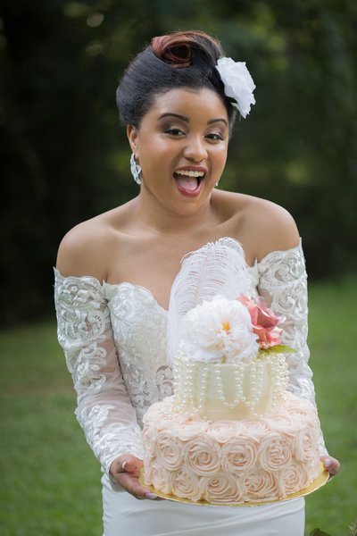 A black bride in a stunning white gown holds a beautifully decorated cake adorned with flowers and pearl details against a lush green background.