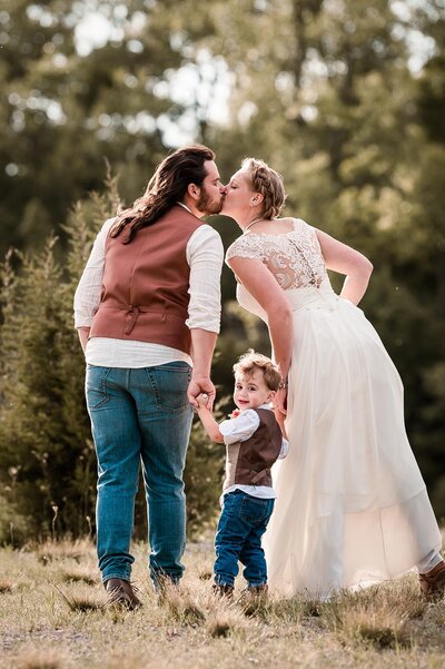 Bride and groom kiss while holding hands with their son and the son looks back at the camera smiling