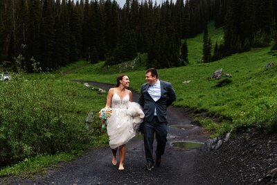 Shannon Lee, a Colorado elopement photographer, captured a newlywed couple joyfully walking on a path through lush green hills. The bride holds a bouquet, and both wear smiles, radiating happiness, love, and the beauty of their newlywed bliss.