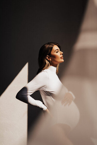 A woman in a white long sleeve bodysuit staring out the side facing the light. In a studio posing holding the top of her belly.
