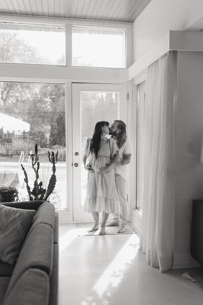A couple shares a loving kiss in their home, celebrating their upcoming arrival in a warm, intimate setting.
