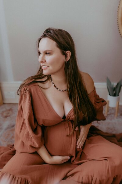 Pregnant woman in a brown dress sits on the floor, cradling her belly with one hand and looking to the side.