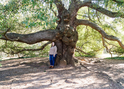 Woman in a tan sweater and jeans standing outdoors surrounded by large twisted tree branches and sunlight filtering through the leaves.