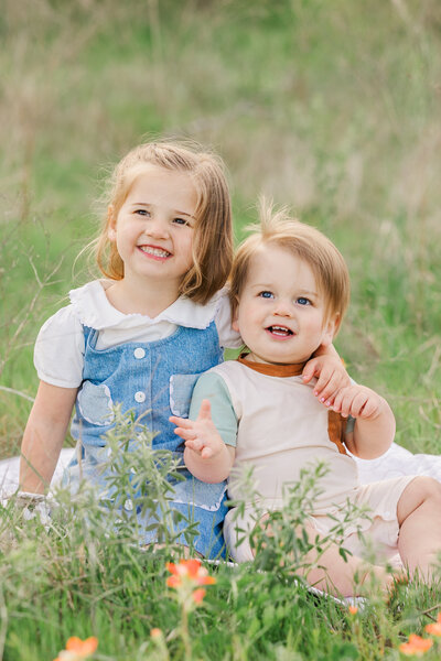Smiling toddler siblings sitting in a field of wildflowers during a spring family photo session in Lewisville, Texas, wearing soft pastel outfits with a natural green backdrop.