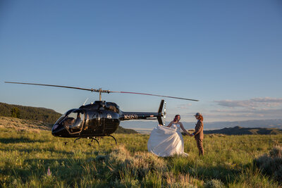 Bride and groom step out of a black helicopter onto an open Montana field during their adventure elopement. The bride's wedding dress flows as they hold hands, surrounded by rolling hills and golden evening light.