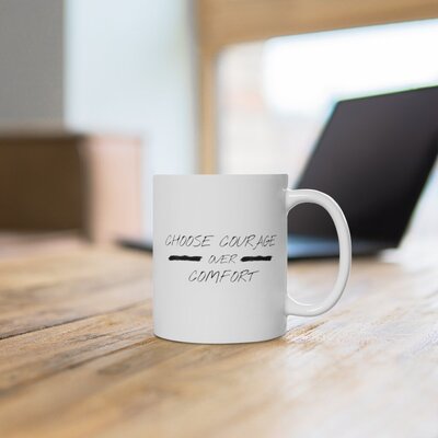 White coffee mug that says "Choose Courage Over Comfort" on a wooden table with a laptop in the background