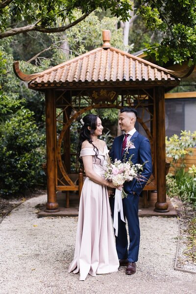 A couple in wedding attire holding a bouquet standing in front of an asian-inspired wooden structure.