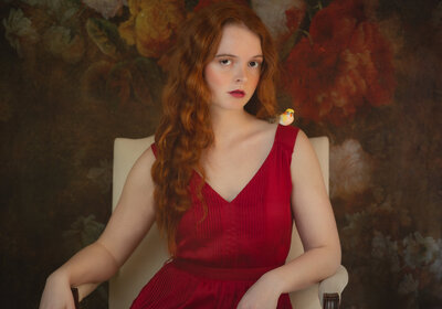 A girl with curly red hair sits in front of an antique backdrop wearing a red dress and has a yellow bird on her shoulder