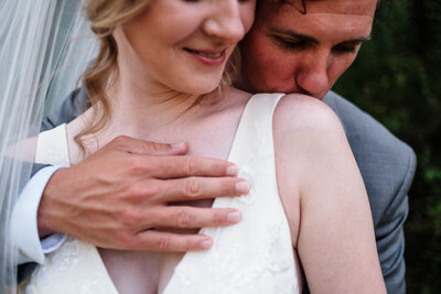 A groom tenderly kisses his bride’s shoulder as she smiles softly, embracing the warmth and intimacy of their wedding day. His hand rests gently over hers, symbolizing love and connection. This heartfelt moment showcases the beauty of Alabama wedding photography.