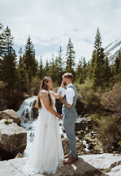 The bride is standing at the base of mountains as she kicks up her leg. She is wearing combat boots and a wedding dress. She has a hiking backpack on with flowers in it. She is smiling towards the ground with one arm lifted in the air. The grass is brown and hte mountains are blue.