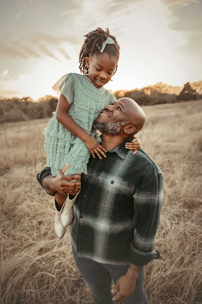 Father and his daughter in our investment page feature. The tall dad, embracing his little girl playfully. The girl, is wearing a mint ruffled dress and a matching bow. The dad is wearing a green plaid shirt and grey pants.They are both standing is a field of tall dry grass.