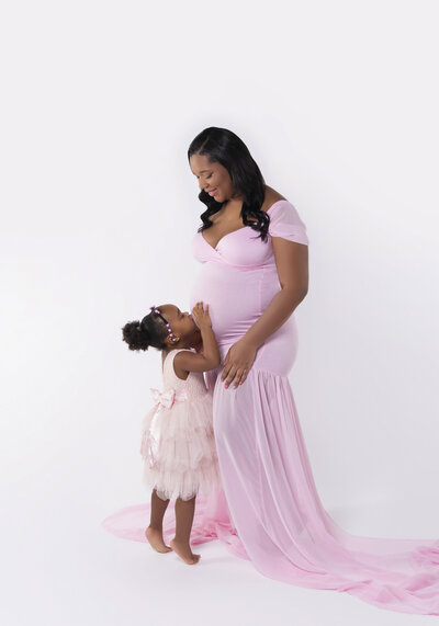 insley Photo expecting Mother and toddler kissing her belly, both wearing pink dresses, with dark hair and dark skin tone. Mom to be is smiling sweetly at her daughter who is standing on her toes to reach up. Created on a white background in our Fort Mill SC portrait studio