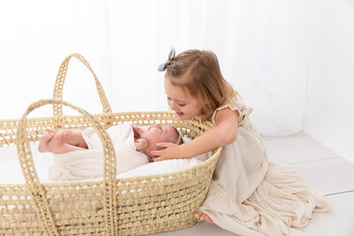 newborn girl wrapped in white fabric in a basket with sister looking at her