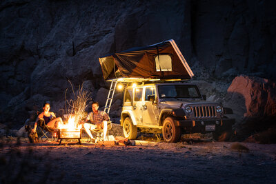 A couple enjoying a private luxury overland camping experience beside a firelit Jeep with a rooftop tent, part of a curated romantic adventure in secluded nature